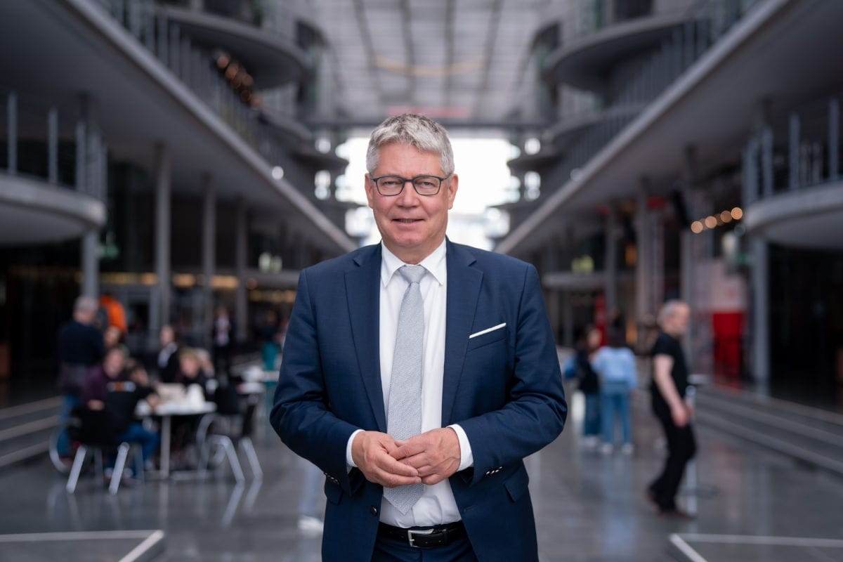 Ein Mann mit grauem Haar und Brille im dunklen Anzug mit Krawatte steht im Fokus vor einer weitläufigen, modernen Architektur mit unscharfen Personen.