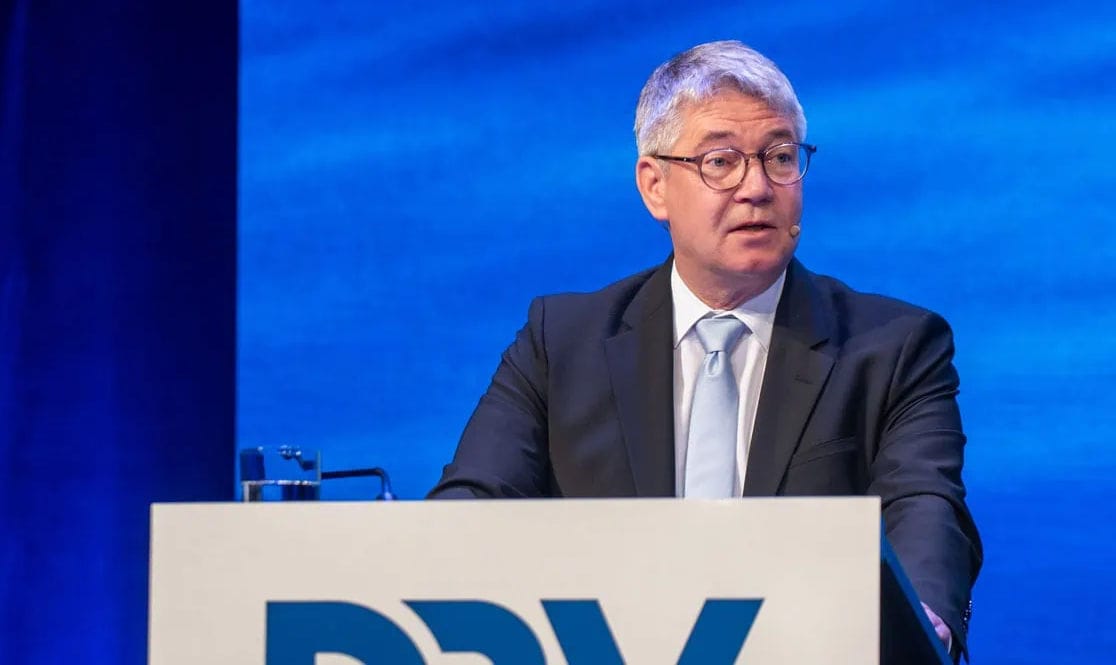 Ein Mann mit grauem Haar und Brille trägt einen dunklen Anzug mit hellblauem Schlips und spricht hinter einem Rednerpult vor einem blauen Hintergrund.
