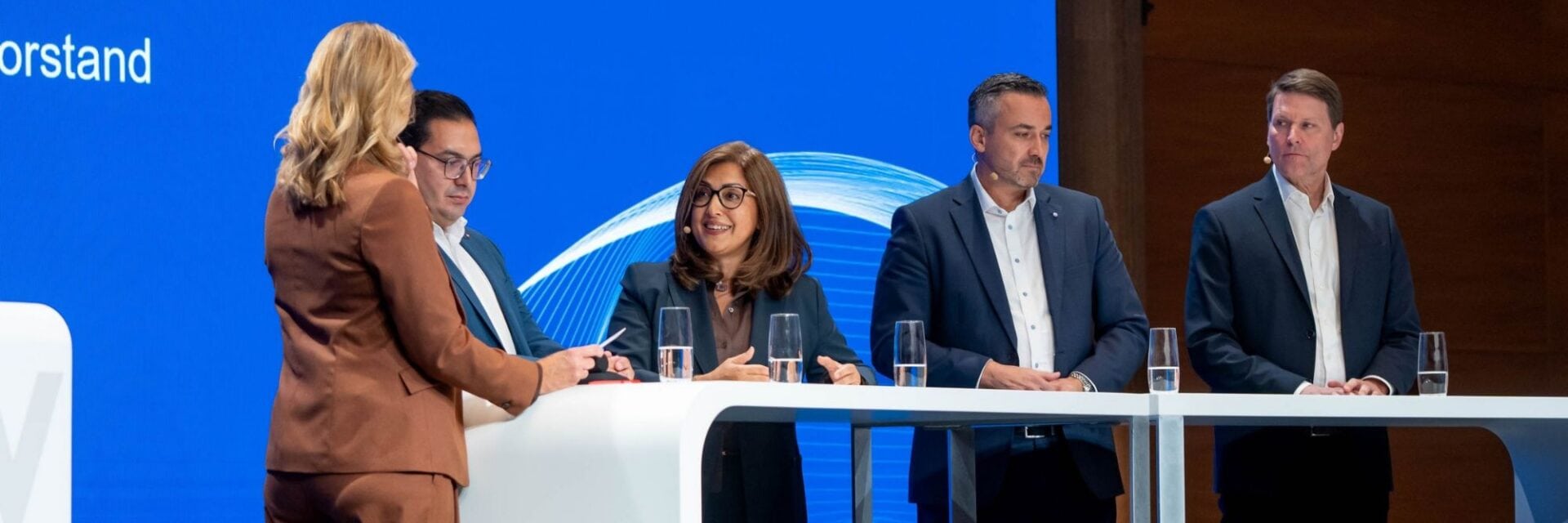 Eine Gruppe von fünf Personen steht hinter einem weißen Konferenztisch vor einer blauen Wand mit grafischen Linien bei einer geschäftlichen Veranstaltung.
