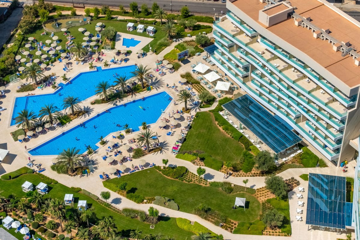 Luftaufnahme eines Hotelkomplexes mit großem, mehrteiligem Außenpool, umgeben von Palmen, Liegeflächen mit Sonnenschirmen und gepflegten Grünanlagen.