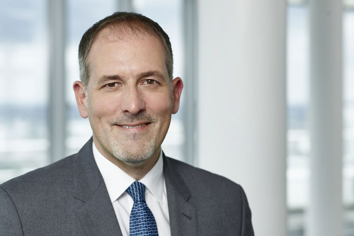 Ein Mann mit kurzem Haar und Kinnbart trägt ein graues Sakko, ein weißes Hemd sowie eine blaue Krawatte und lächelt freundlich in die Kamera vor hellem Grund.