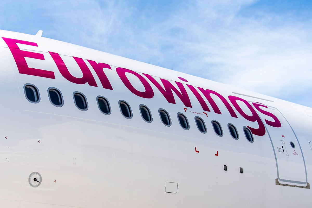 Nahaufnahme des weißen Rumpfes eines Eurowings-Flugzeugs mit dem markanten magentafarbenen Logo und einer Reihe von Kabinenfenstern vor blauem Himmel.