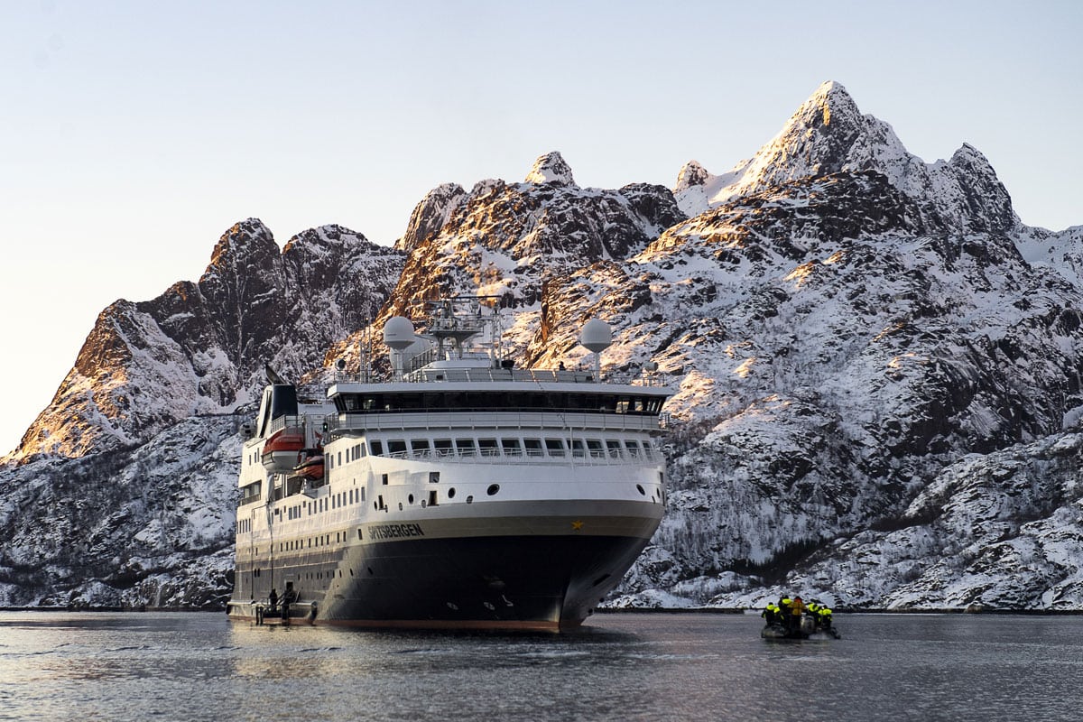 Das Kreuzfahrtschiff Spitsbergen liegt vor einer schneebedeckten Bergkulisse im Wasser, während ein kleines Schlauchboot mit Personen daneben manövriert.