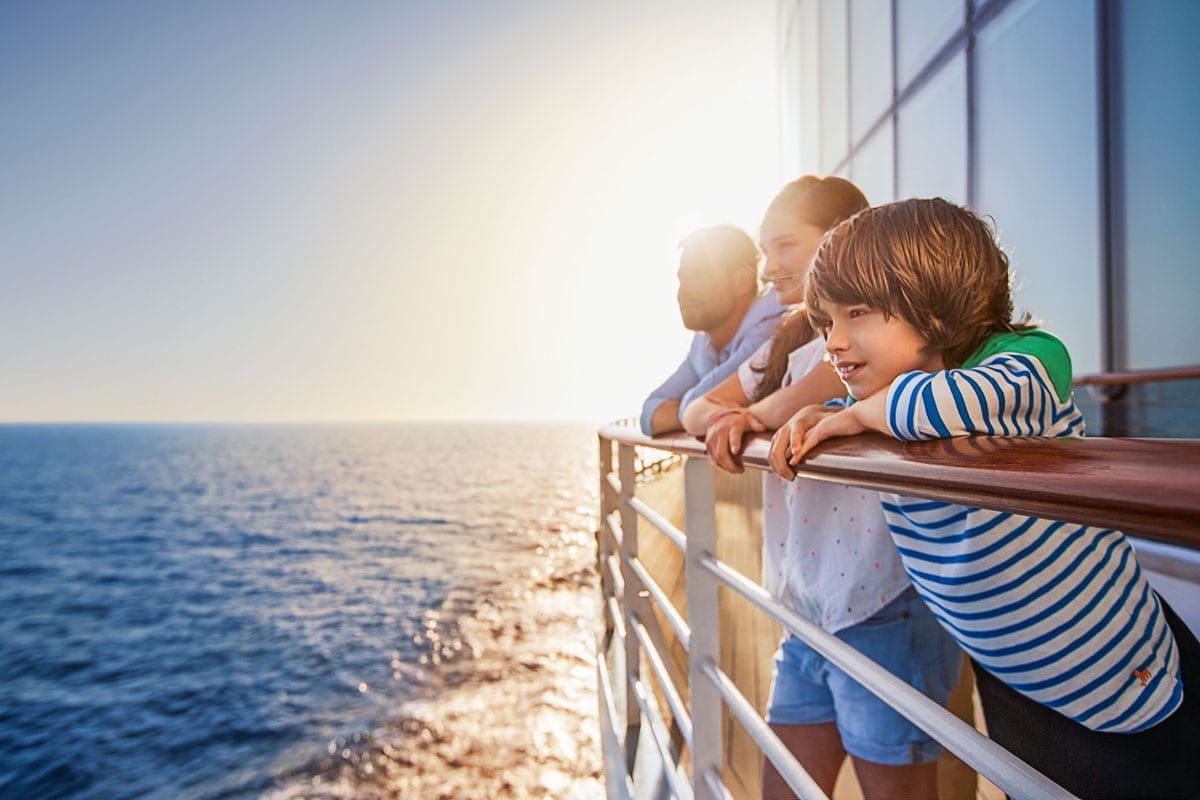 Ein Mann, eine Frau und ein Kind stehen an der Reling eines Kreuzfahrtschiffes und blicken gemeinsam auf das offene Meer während des hellen Sonnenuntergangs.