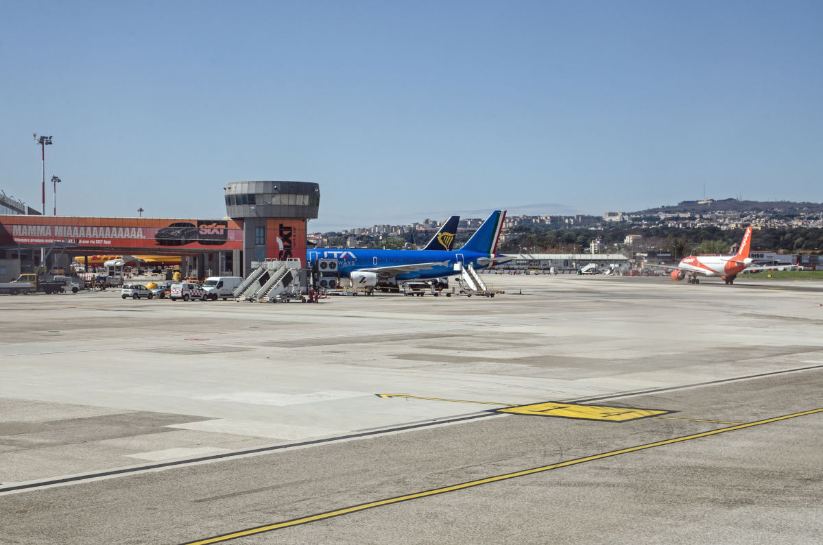 Blick auf ein sonniges Flughafenvorfeld mit mehreren Passagierflugzeugen, einem Kontrollturm und Bodenabfertigungsfahrzeugen unter einem klaren blauen Himmel.