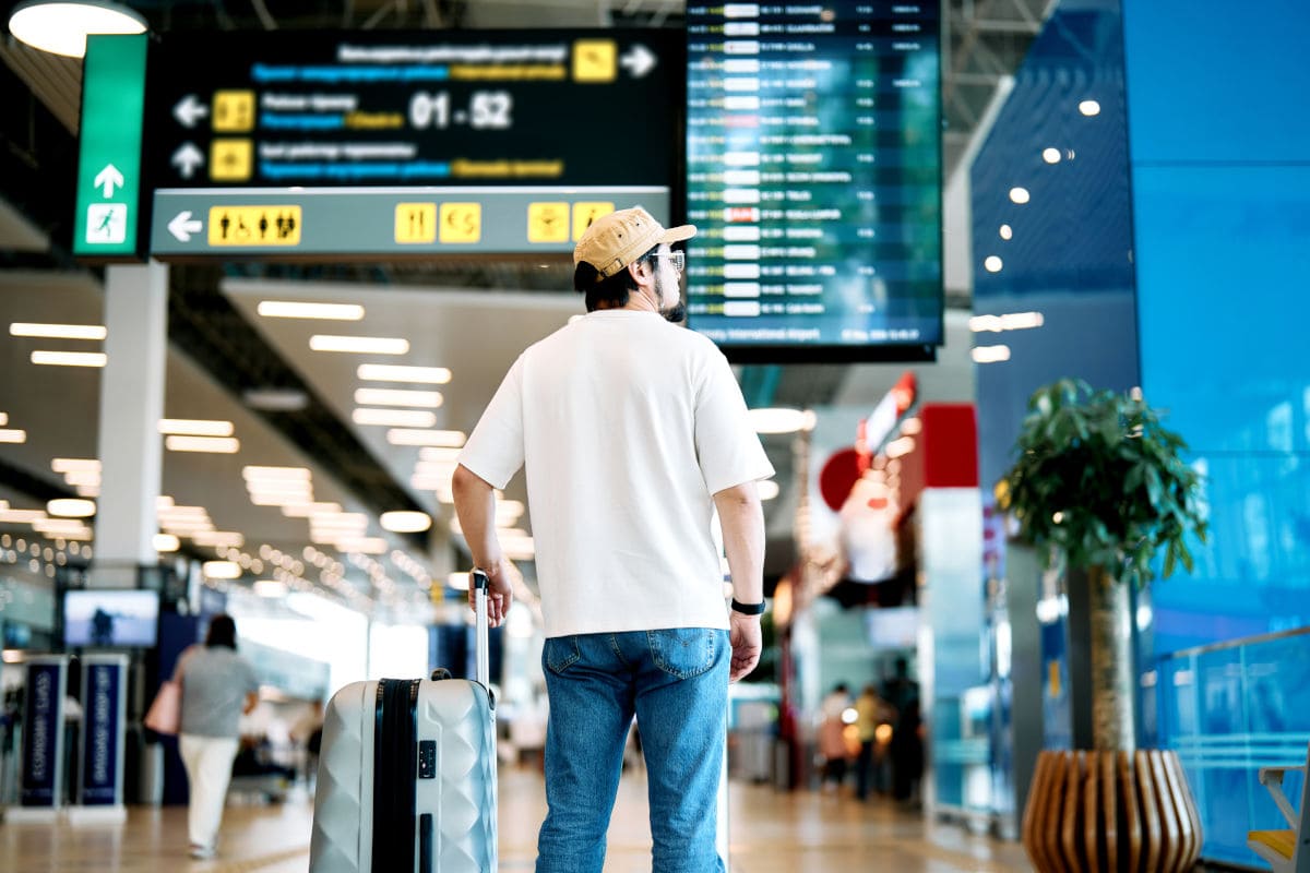 Ein Mann in weißem T-Shirt, Jeans und beiger Kappe steht mit einem Rollkoffer in einer Flughafenhalle und blickt auf eine große digitale Anzeigetafel.