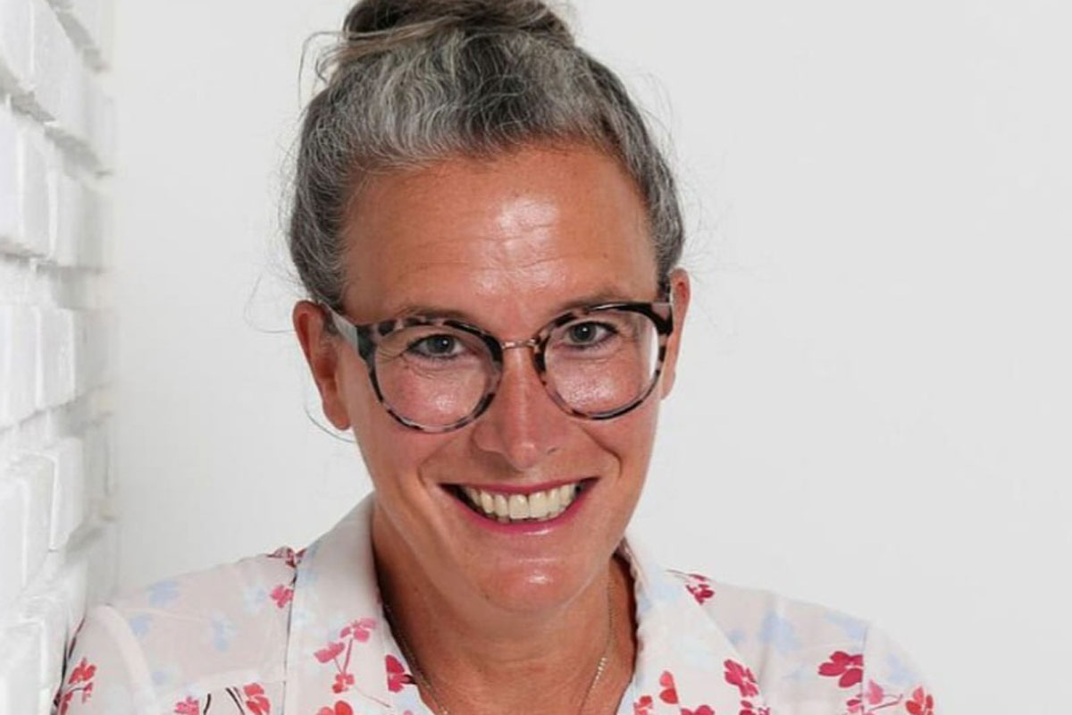 Eine lächelnde Frau mit grauem Haar im Dutt und einer gemusterten Brille trägt eine weiße Bluse mit floralem Muster vor einer weißen Backsteinwand.
