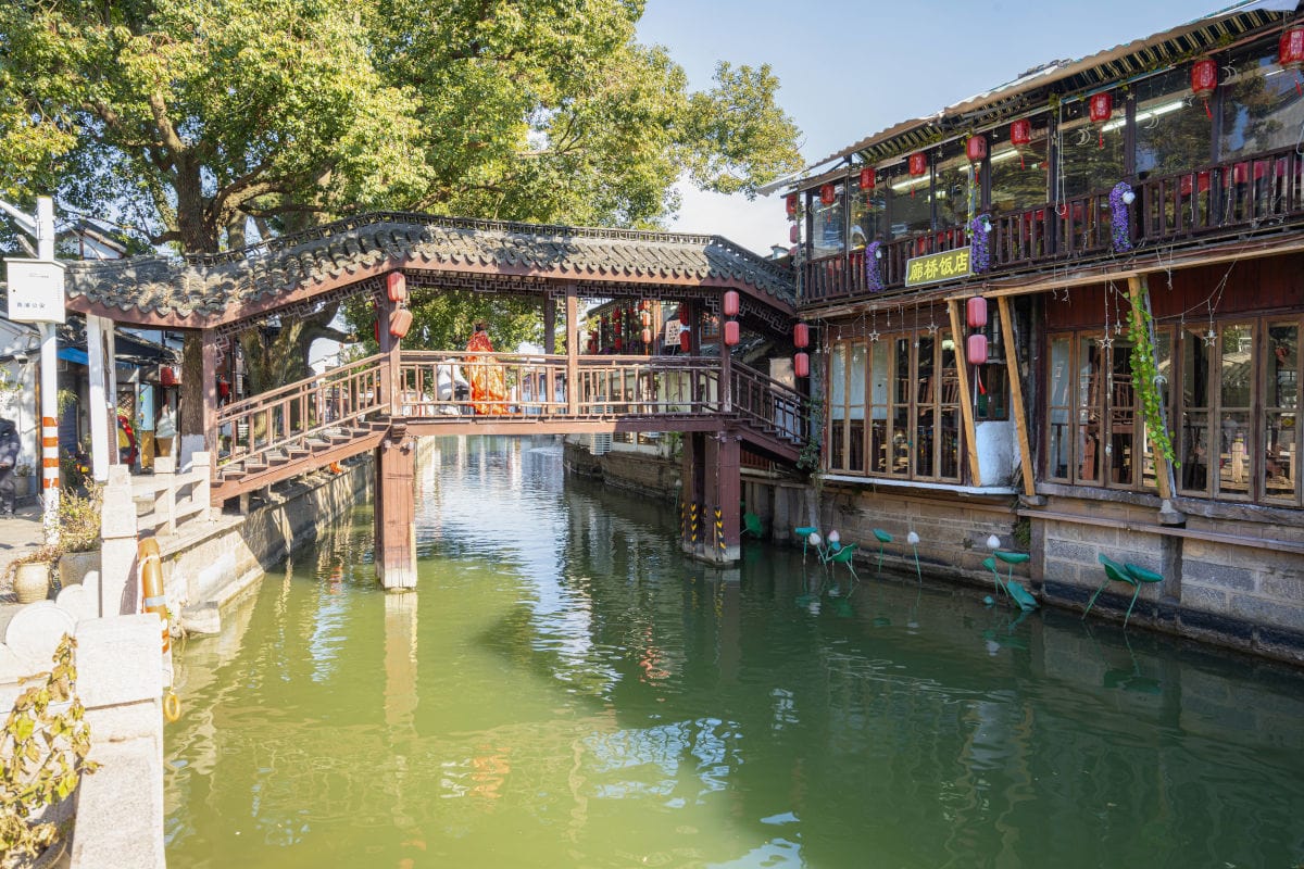 Eine überdachte Holzbrücke führt über einen Kanal in einer historischen chinesischen Wasserstadt, flankiert von traditionellen Gebäuden mit roten Lampions.