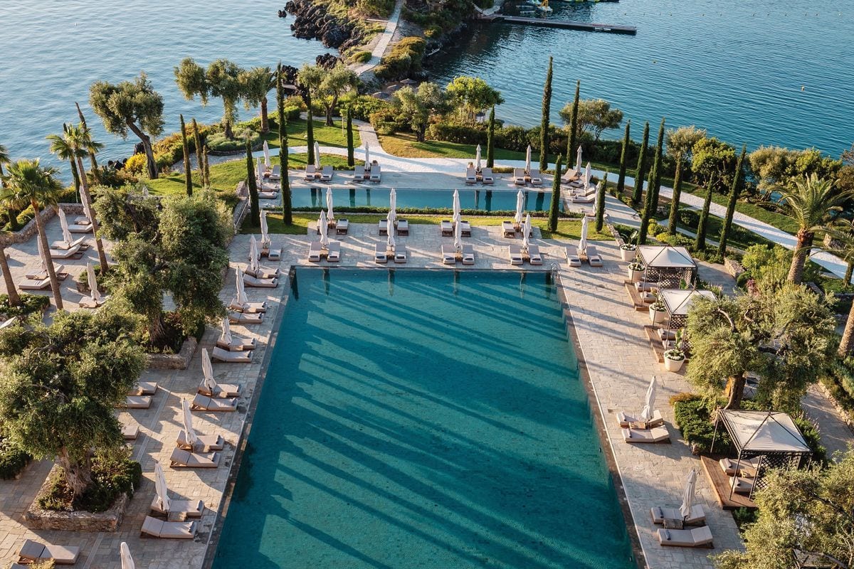 Luftaufnahme einer Hotelanlage mit zwei rechteckigen Swimmingpools, umgeben von Liegestühlen, Sonnenschirmen, Olivenbäumen und Zypressen direkt am Meer.