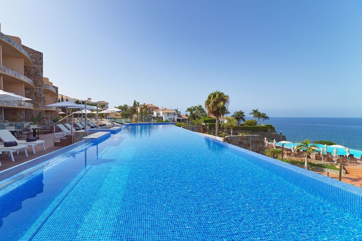 Ein langer Infinity-Pool mit blauem Mosaik erstreckt sich vor einem Hotelgebäude mit Terrassen, Palmen und einem weiten Blick auf das offene, blaue Meer.