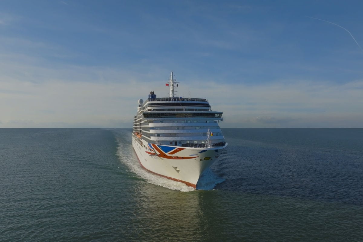 Ein weißes Kreuzfahrtschiff der Reederei P&O Cruises fährt frontal auf das offene Meer hinaus, während es eine deutliche Bugwelle im ruhigen Wasser erzeugt.