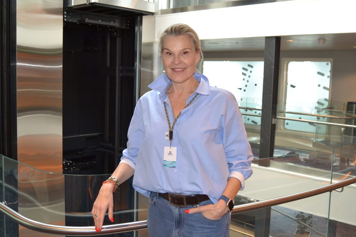 Eine blonde Frau in einem hellblauen Hemd und Jeans steht lächelnd vor einem gläsernen Aufzug in einem modernen, hellen Gebäude mit Glasgeländer und Fenstern.