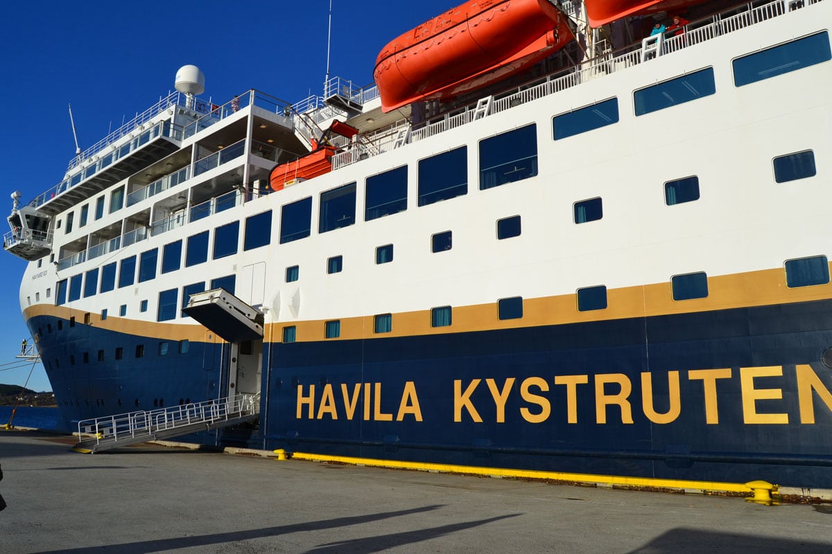 Die Seitenansicht eines Havila Kystruten Schiffes an einem Kai mit ausgefahrener Gangway und Rettungsbooten vor einem klaren, blauen Himmel bei Tageslicht.