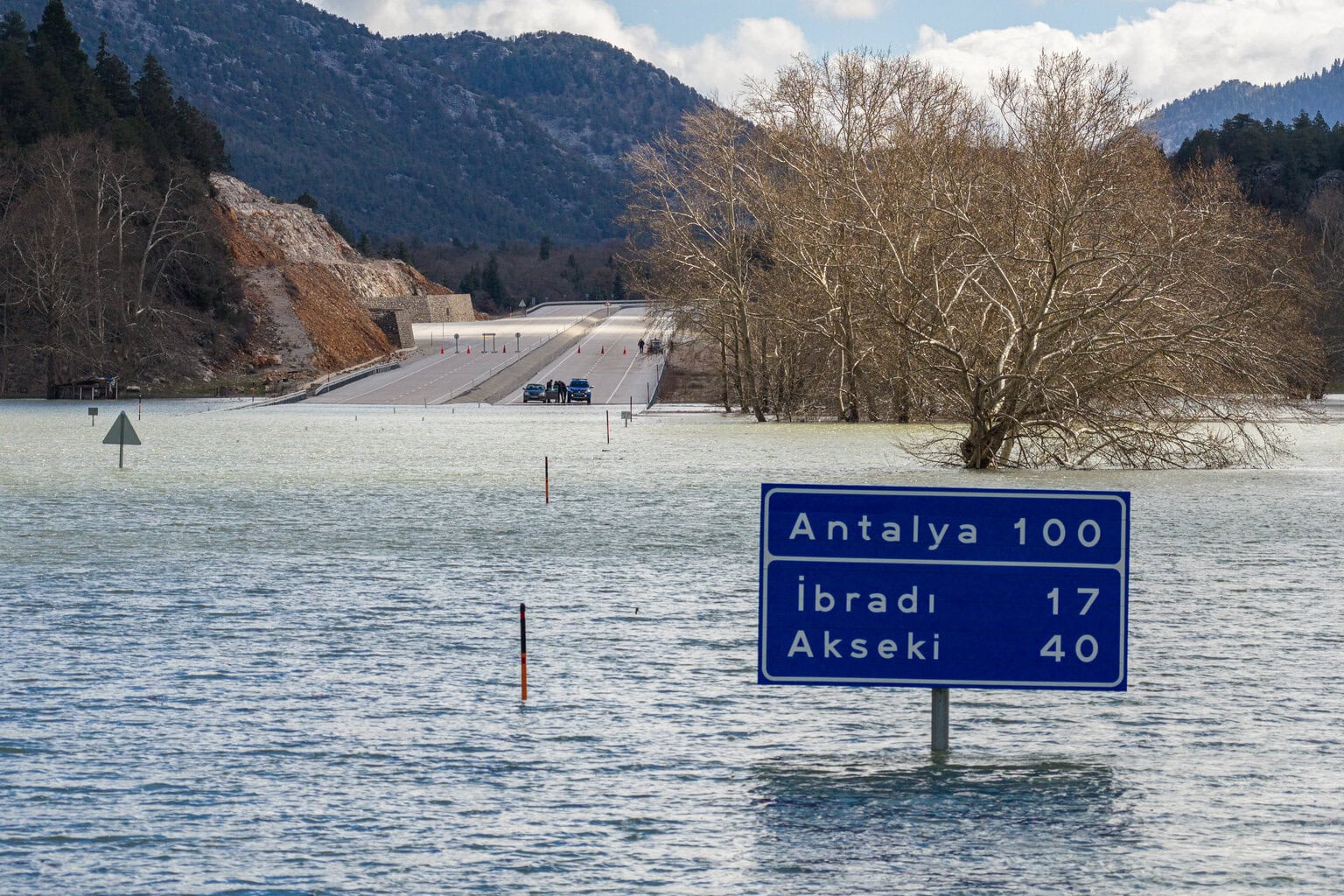Ein blaues Straßenschild mit Entfernungsangaben nach Antalya, İbradı und Akseki steht in einer überfluteten Landschaft vor einer unterbrochenen Fahrbahn.