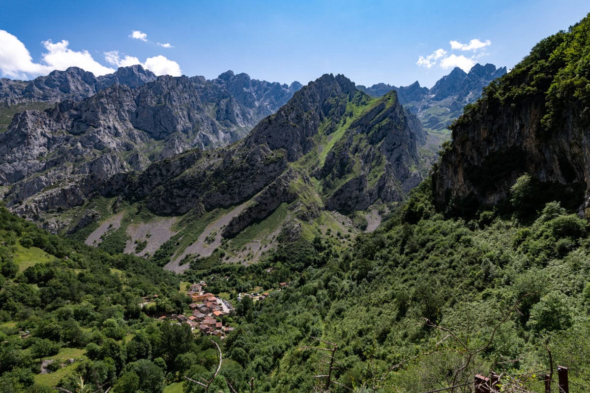 Ein kleines Dorf liegt in einem tiefen, grünen Tal, umgeben von steilen, schroffen Felswänden und hohen, zerklüfteten Berggipfeln unter einem blauen Himmel.