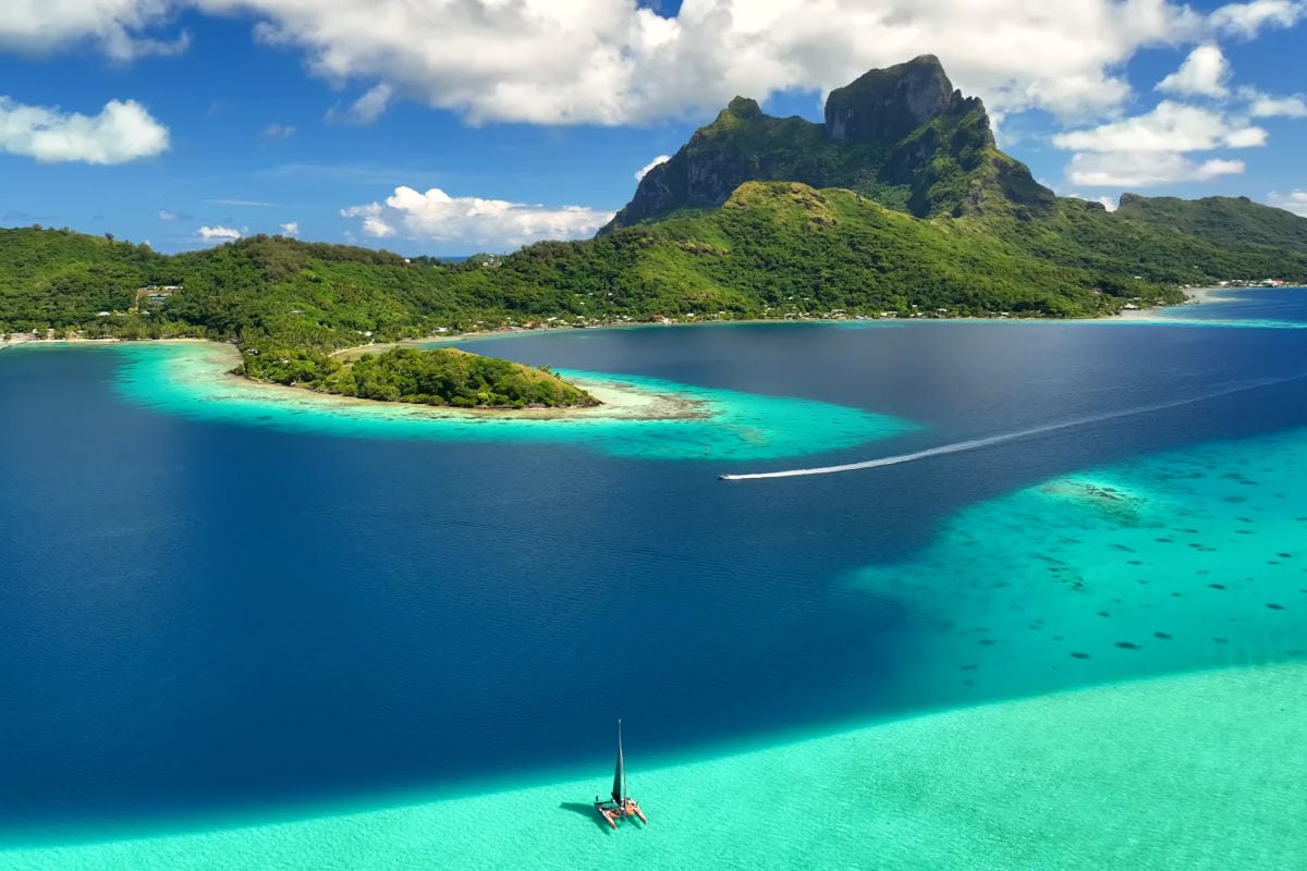 Luftaufnahme einer tropischen Lagune mit türkisfarbenem Wasser, einem Segelboot im Vordergrund, einer kleinen Insel und einem markanten Berg im Hintergrund.