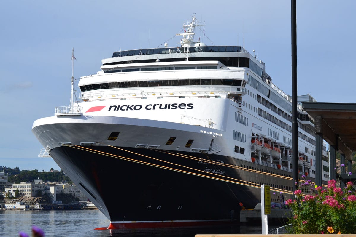Das Kreuzfahrtschiff Vasco da Gama von nicko cruises liegt an einem Kai vor einer Küstenlandschaft, im Vordergrund sind unscharfe Blumen zu sehen.