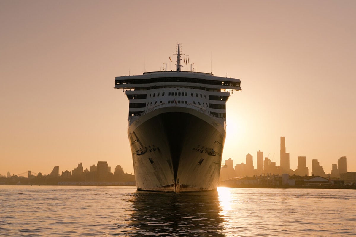 Die Queen Mary 2 fährt frontal auf den Betrachter zu, während im Hintergrund bei warmem Sonnenuntergang die Skyline einer Großstadt am Wasser zu sehen ist.