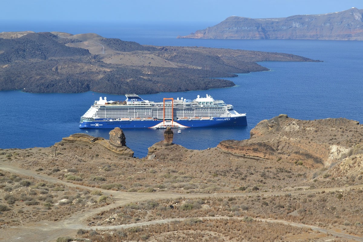 Ein großes Kreuzfahrtschiff der Celebrity Apex liegt vor der Küste einer kargen, felsigen Inselgruppe im tiefblauen Meer bei klarem, sonnigem Tageslicht.