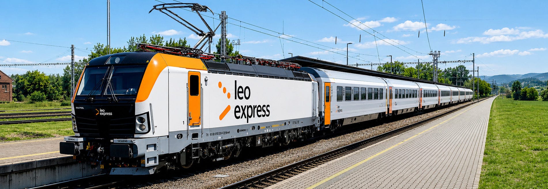 leo-express-frankfurt-streichung Ein weiß-orangefarbener Leo Express Zug steht an einem Bahnsteig in einer ländlichen Umgebung unter blauem Himmel mit Oberleitungen im Hintergrund.