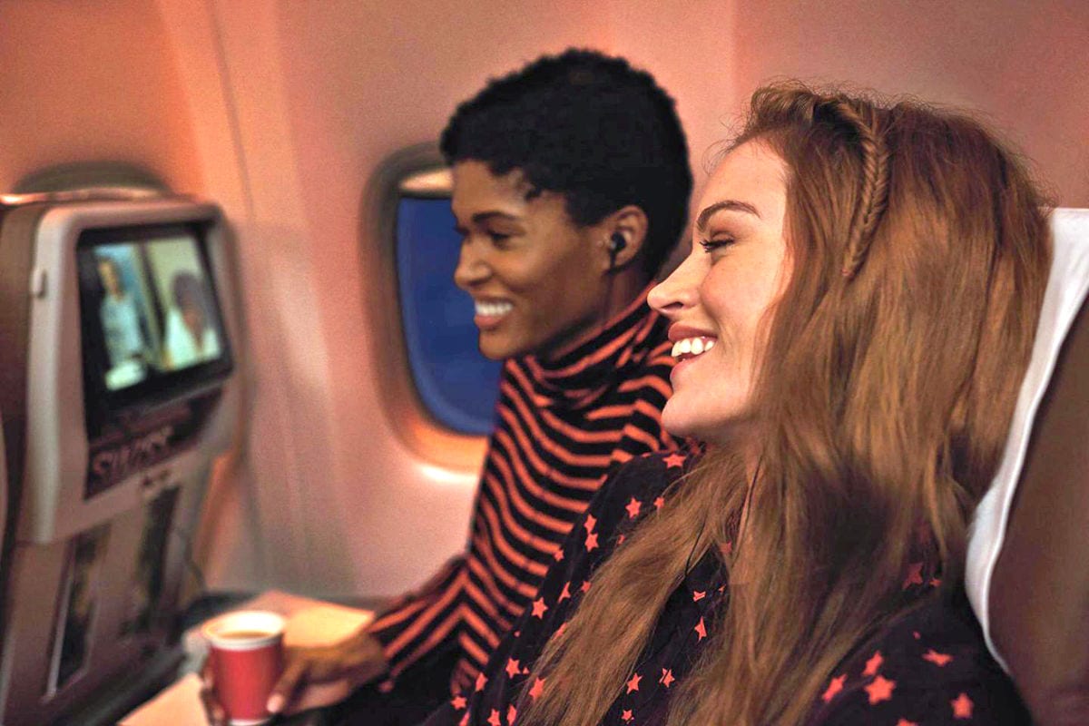 Zwei junge Frauen sitzen nebeneinander in einem Flugzeugsitz, schauen lächelnd auf einen Bildschirm vor ihnen und tragen Kopfhörer in einer Kabinenumgebung.