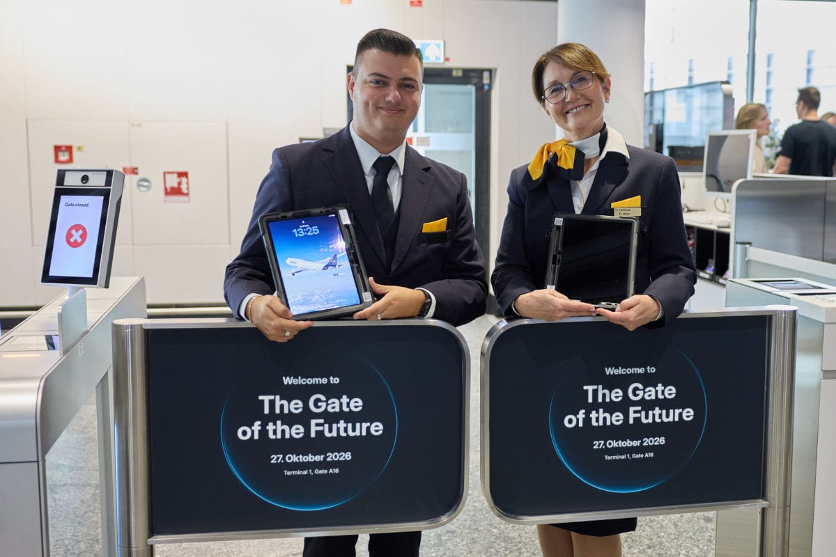 Ein Mann und eine Frau in dunklen Uniformen stehen an einem Flughafengate und halten Tablets vor sich, während sie in die Kamera lächeln und posieren.