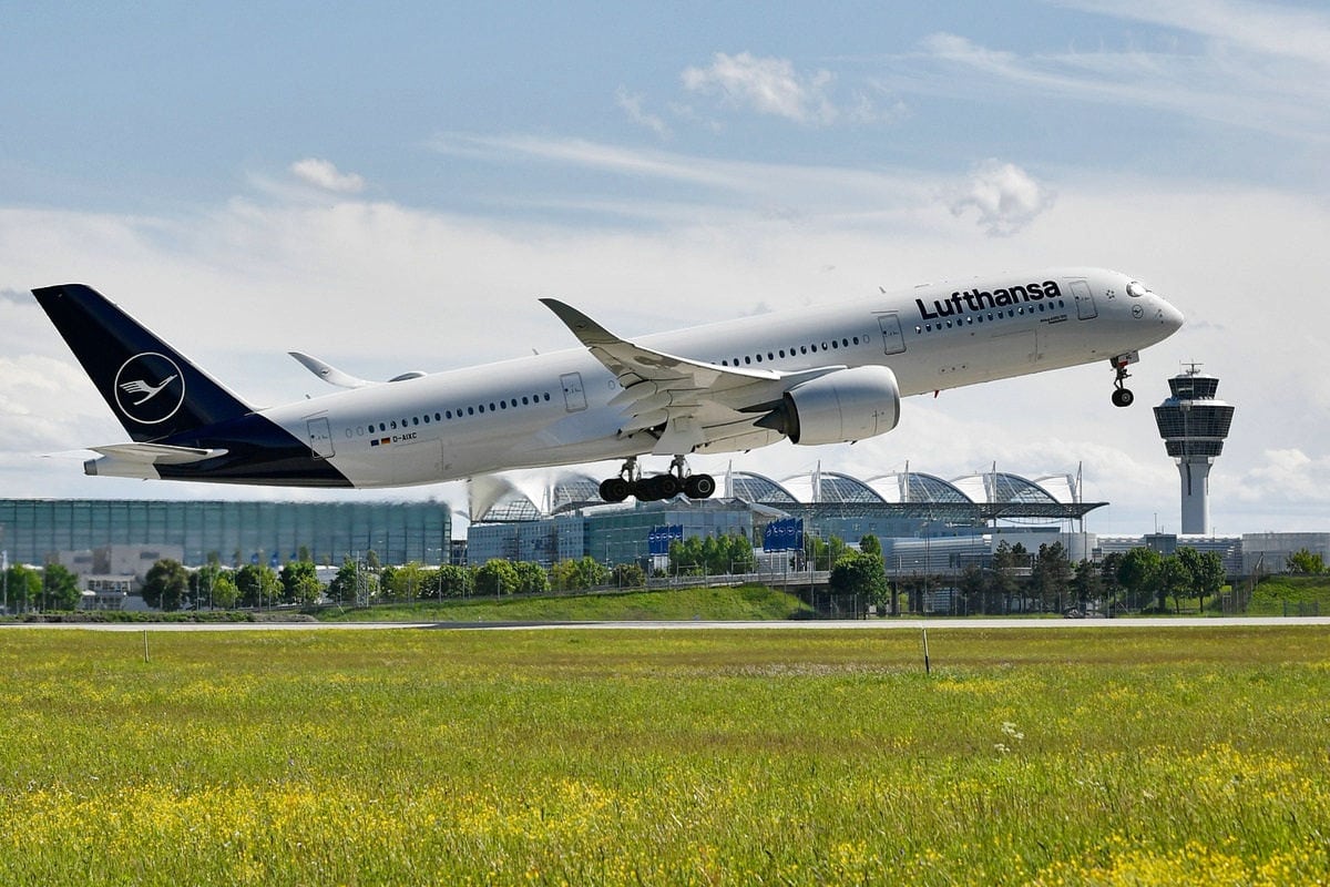 Ein Lufthansa-Passagierflugzeug vom Typ Airbus A350 hebt von einer Startbahn ab, im Hintergrund sind der Tower und Flughafengebäude bei Tageslicht zu sehen.