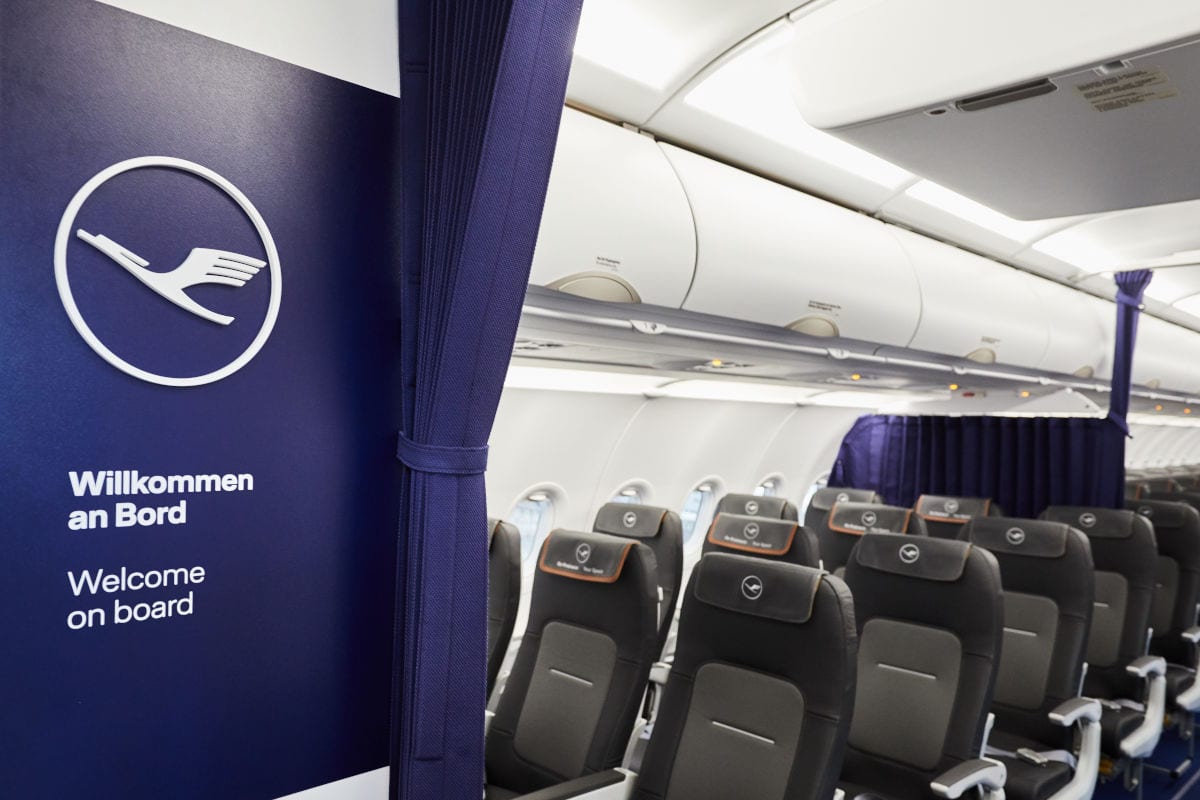 Blick in die Flugzeugkabine mit Lufthansa-Logo und Willkommensgruß auf einer dunkelblauen Trennwand sowie Reihen von grauen Passagiersitzen im Hintergrund.