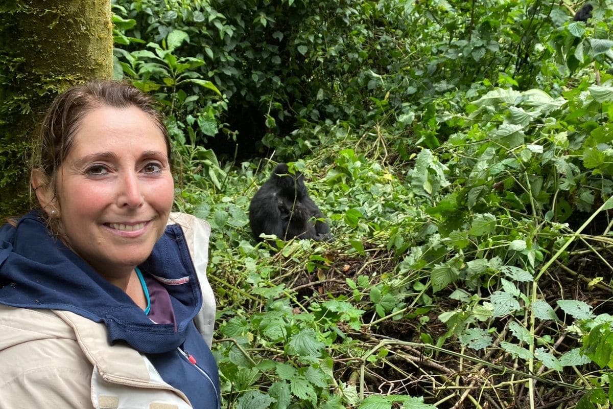 Eine lächelnde Frau im Vordergrund und ein Gorilla im Hintergrund, der inmitten von dichtem, grünem Unterholz in einem Waldgebiet sitzt und frisst.