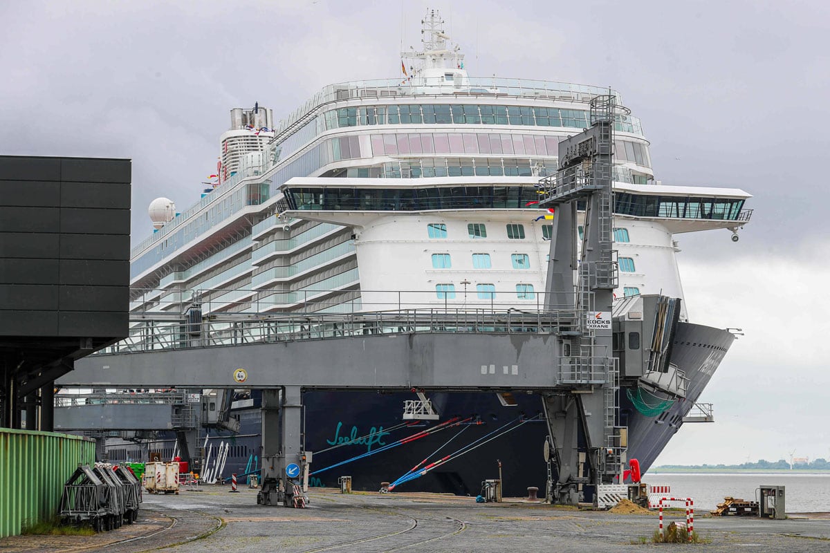 Ein großes Kreuzfahrtschiff liegt an einem Kai im Hafen. Im Vordergrund befindet sich eine graue Passagierbrücke, die mit dem Schiff verbunden ist.