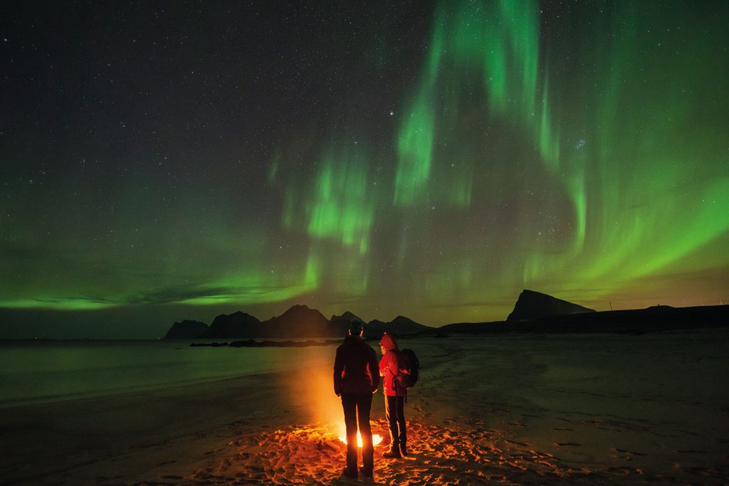 Zwei Personen stehen bei Nacht an einem Lagerfeuer an einem Sandstrand, während am dunklen Himmel über den Bergen leuchtend grüne Polarlichter zu sehen sind.