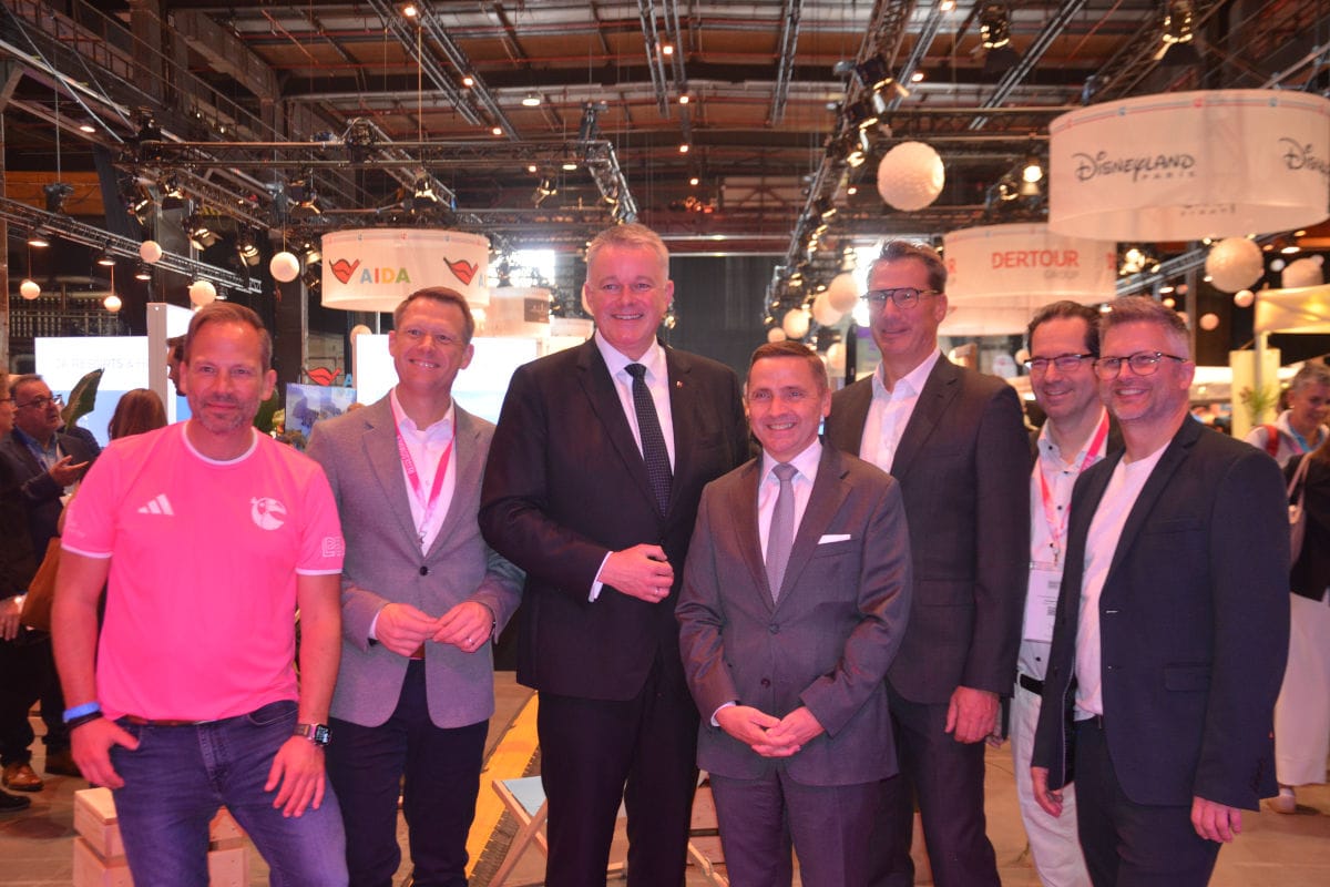 Eine Gruppe von sieben Männern steht in einer Messehalle vor verschiedenen Firmenlogos wie AIDA und Dertour für ein gemeinsames Foto nebeneinander.