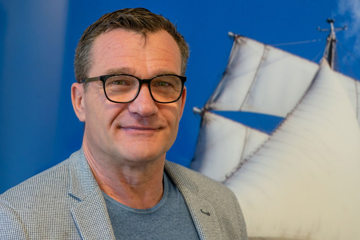 Ein Mann mit Brille und grauem Sakko blickt in die Kamera, im Hintergrund ist ein großes Wandbild mit den weißen Segeln eines Schiffes vor blauem Himmel.
