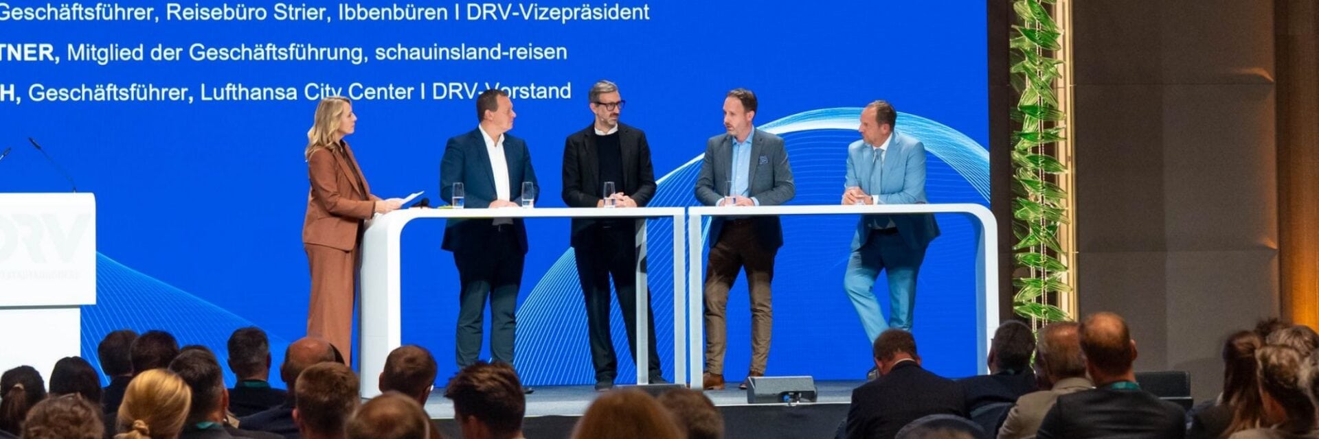 reisebuero-umsaetze Eine Frau und vier Männer stehen auf einer Bühne vor einer blauen Leinwand mit Text, während das Publikum im Vordergrund den Vortrag aufmerksam verfolgt.