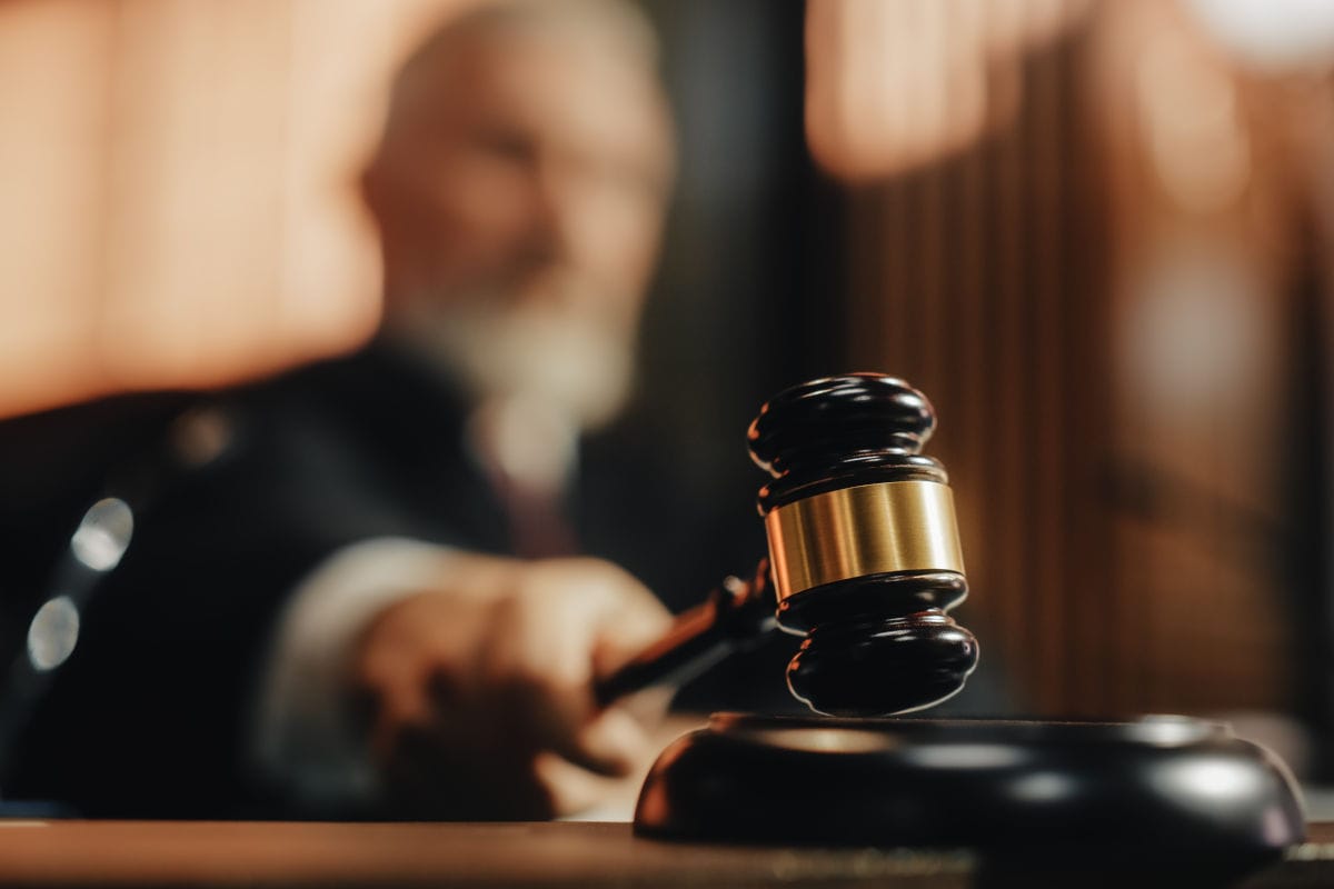 Ein hölzerner Auktionshammer mit goldenem Ring schlägt auf einen Resonanzboden, während im unscharfen Hintergrund eine Person in dunkler Kleidung sitzt.