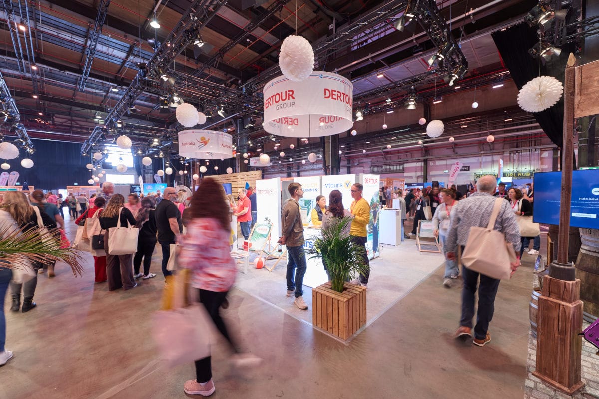 Blick in eine große Messehalle mit Ständen der DERTOUR Group, vielen Besuchern, weißen Papierlampions an der Decke und einer Topfpflanze im Vordergrund.