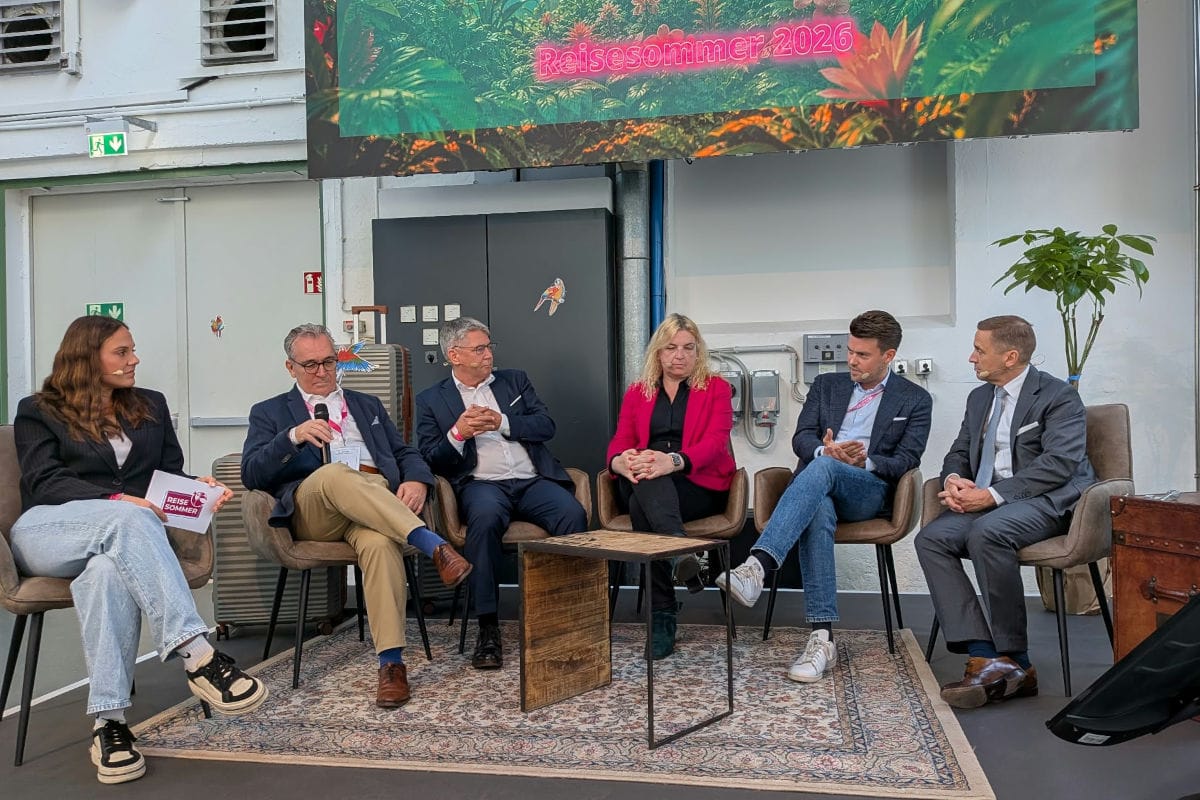 Sechs Personen sitzen in einer Reihe auf Sesseln auf einer Bühne vor einem Bildschirm mit der Aufschrift Reisesommer 2026 und diskutieren miteinander.
