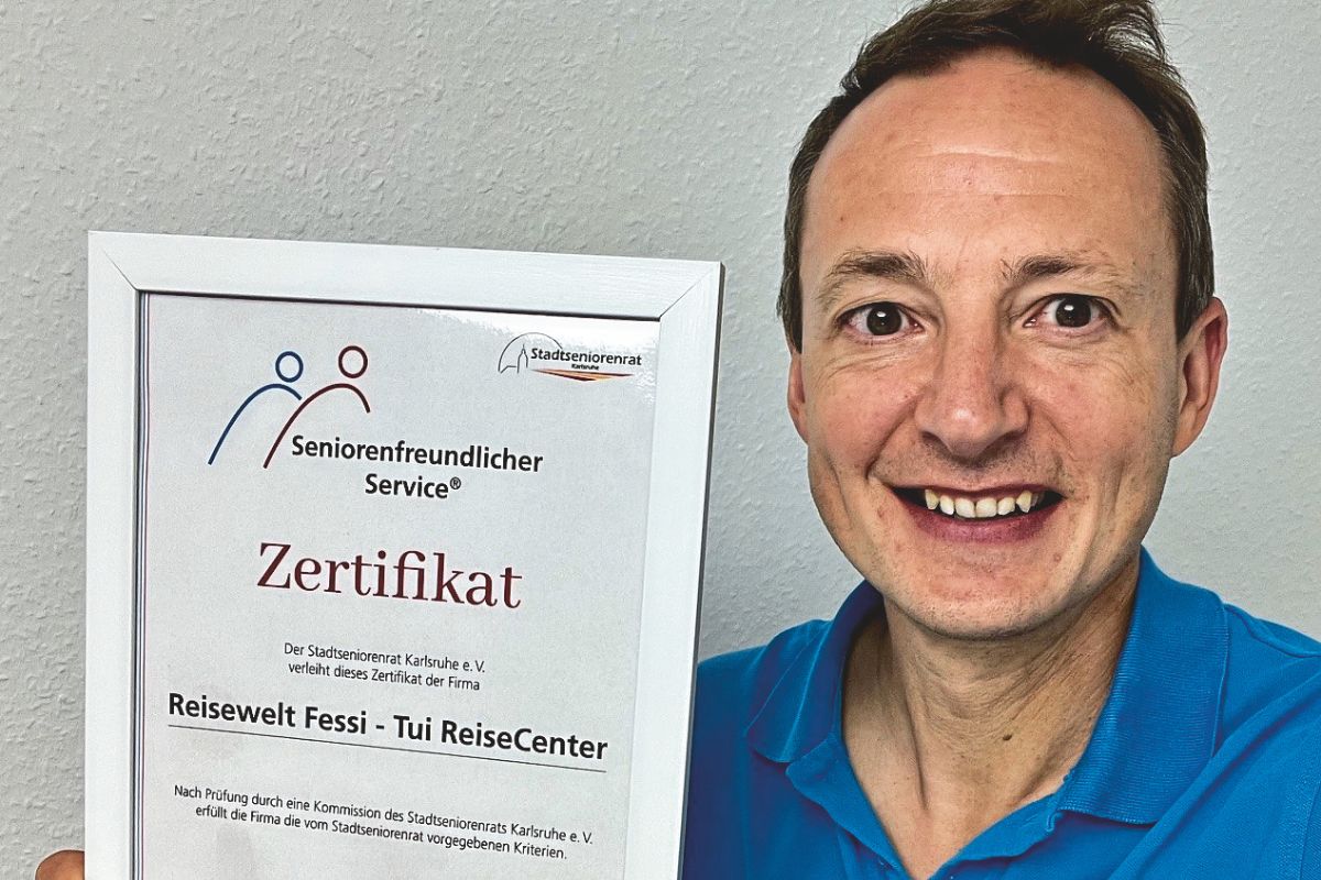 Ein lächelnder Mann in einem blauen Poloshirt hält ein gerahmtes Zertifikat für seniorenfreundlichen Service vom Stadtseniorenrat Karlsruhe in die Kamera.