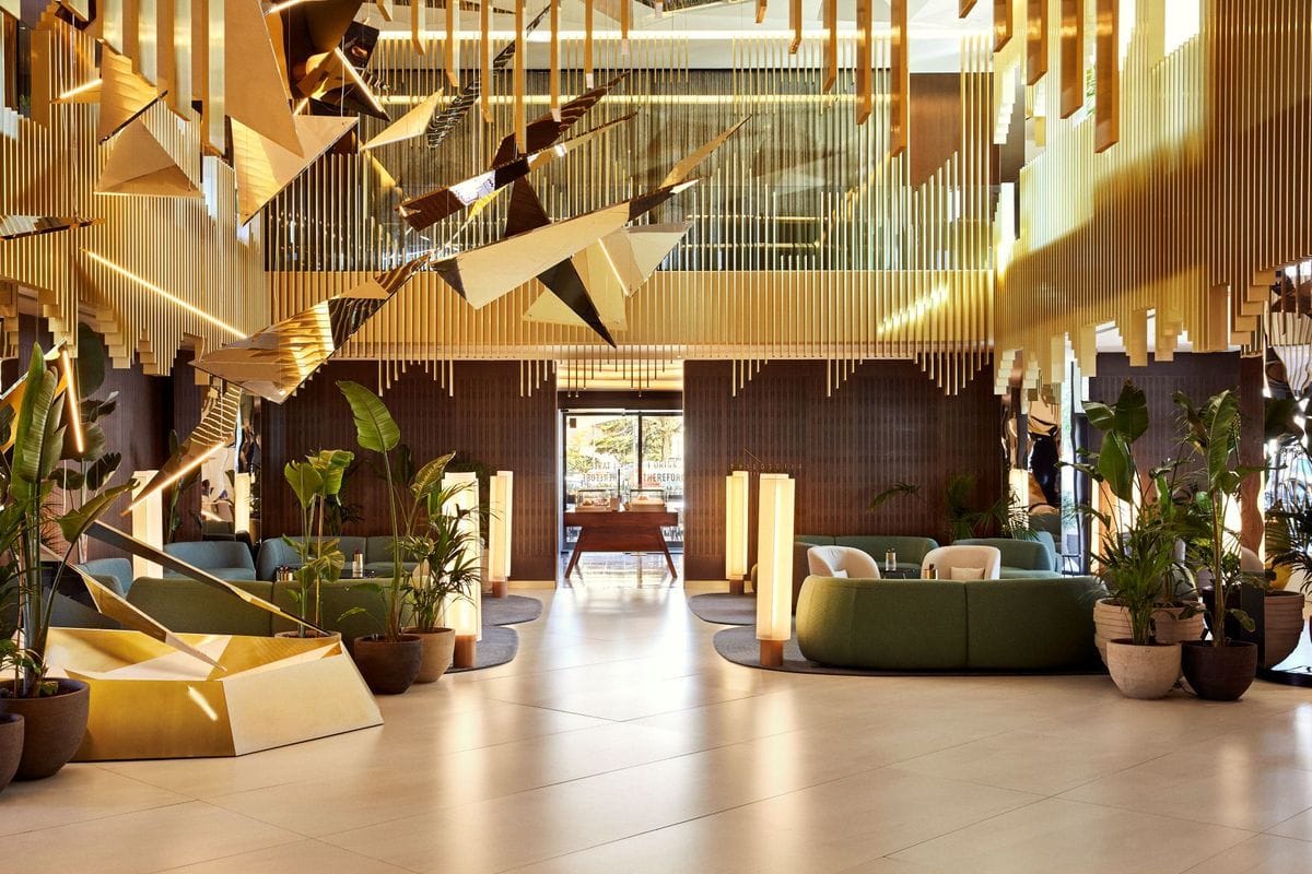 Die weitläufige Hotellobby zeigt eine goldene Deckeninstallation, grüne Sitzgruppen, Topfpflanzen und eine zentrale Durchsicht zum hellen Eingangsbereich.