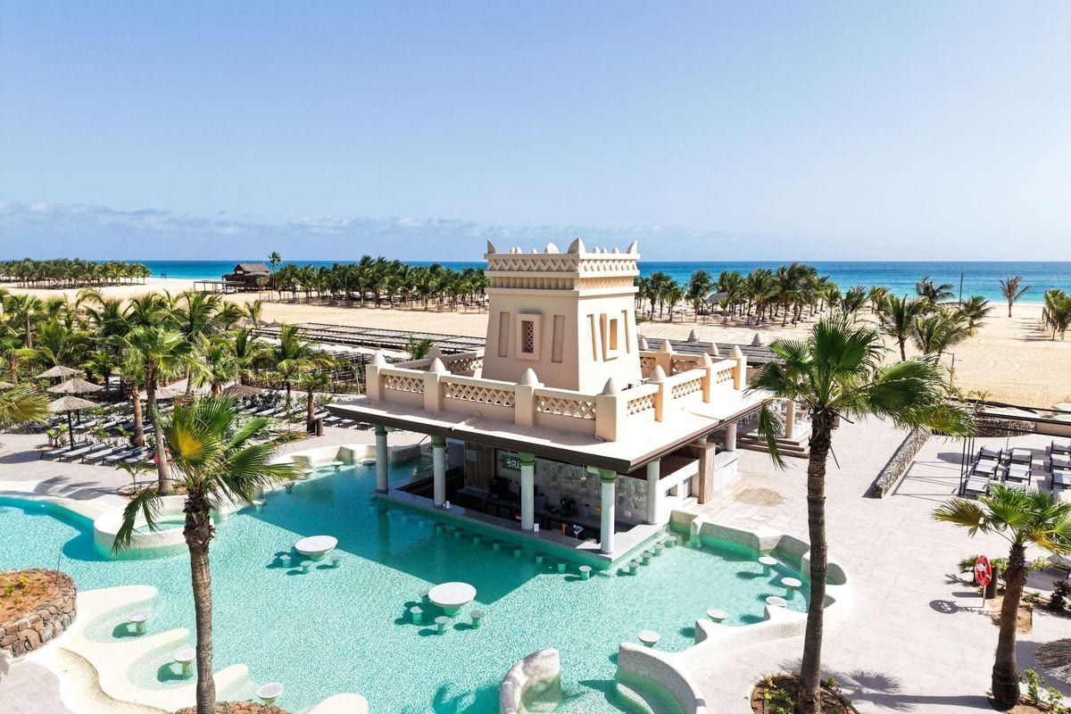 Blick von oben auf eine Poolbar mit turmartigem Gebäude, umgeben von Palmen, einem hellen Sandstrand und dem blauen Meer unter einem klaren Himmel.