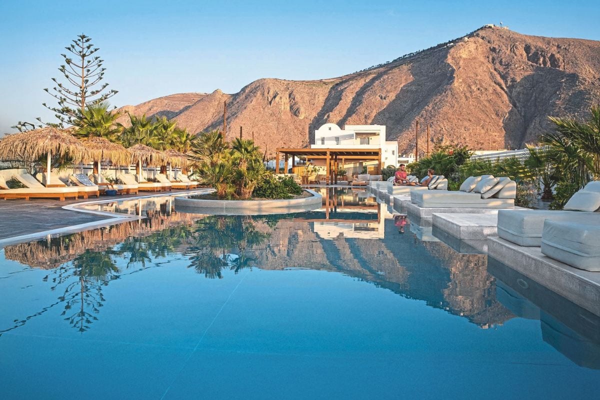 Ein großer Swimmingpool mit spiegelnder Wasseroberfläche, umgeben von Sonnenliegen, Sonnenschirmen und Palmen vor einer imposanten Bergkulisse bei Tageslicht.