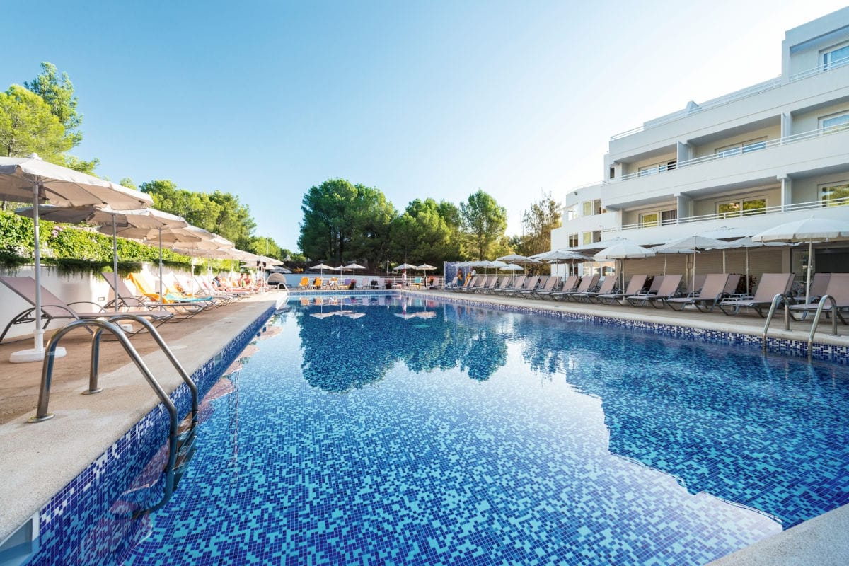 Ein langer, rechteckiger Hotelpool mit blauem Mosaikfliesenbelag, umgeben von zahlreichen Sonnenliegen und Sonnenschirmen vor einem weißen Hotelgebäude.