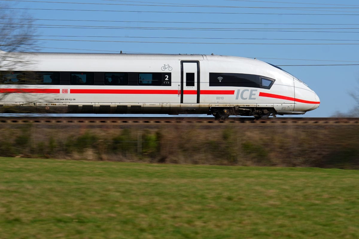 Ein weißer ICE der Deutschen Bahn fährt in hoher Geschwindigkeit auf einer Strecke an einer grünen Wiese und kahlen Bäumen vor einem blauen Himmel vorbei.