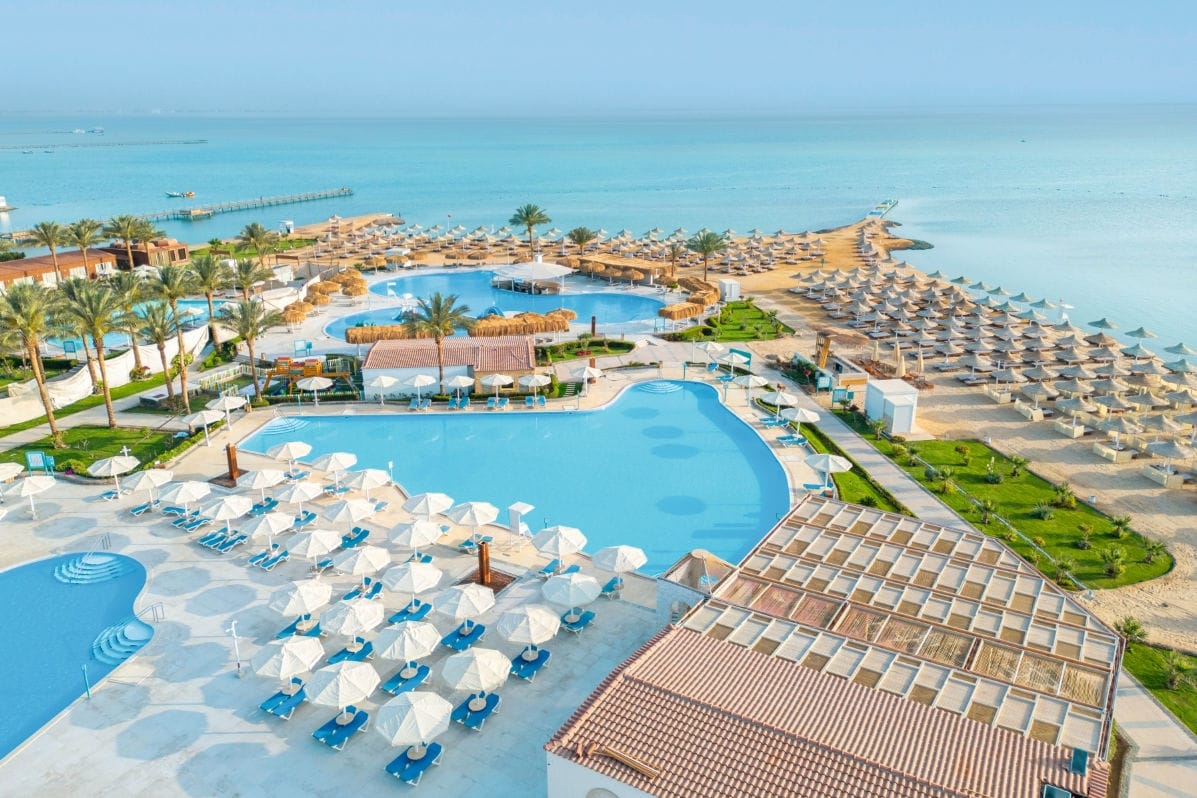 Luftaufnahme einer Hotelanlage mit mehreren großen, hellblauen Swimmingpools, zahlreichen weißen Sonnenschirmen, Liegen und direktem Zugang zum Meer.