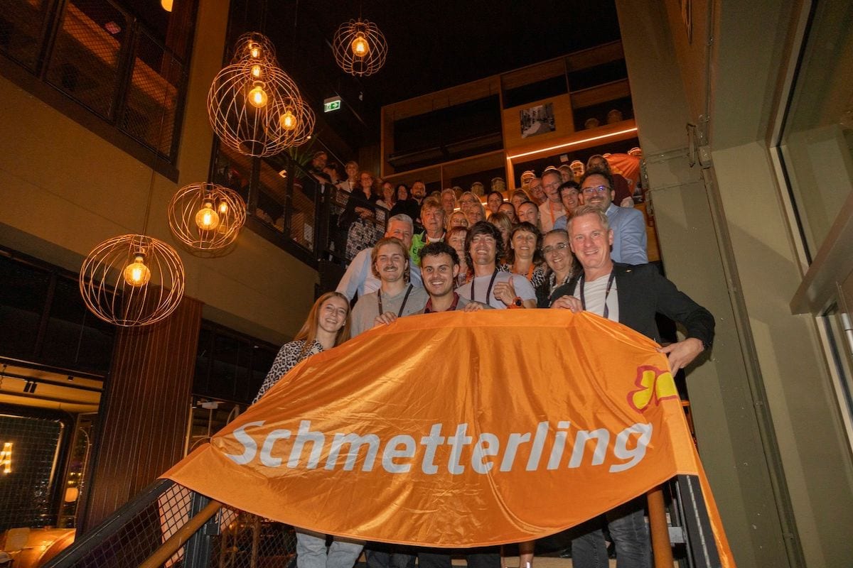 Eine große Gruppe von Menschen steht auf einer Treppe in einem modern beleuchteten Innenraum und hält gemeinsam eine orangefarbene Schmetterling-Fahne.