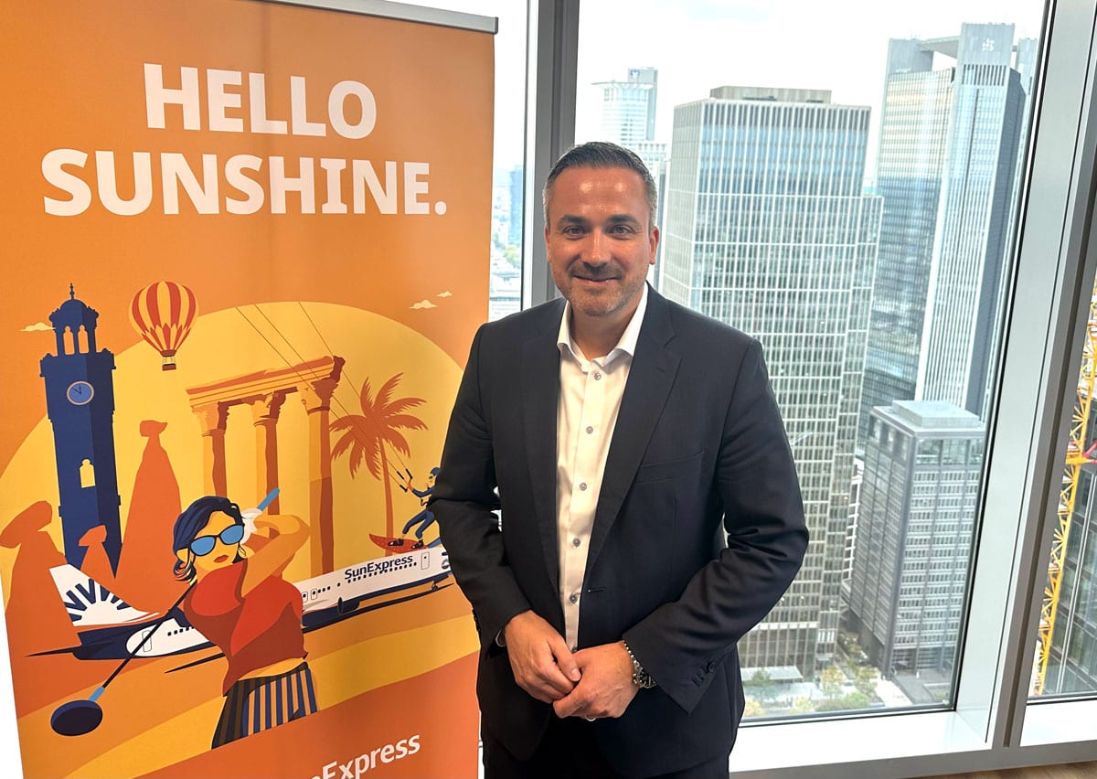 Ein Mann im dunklen Anzug und weißem Hemd steht vor einem orangefarbenen Roll-up-Banner mit Reise-Illustrationen und Blick auf eine moderne Skyline.