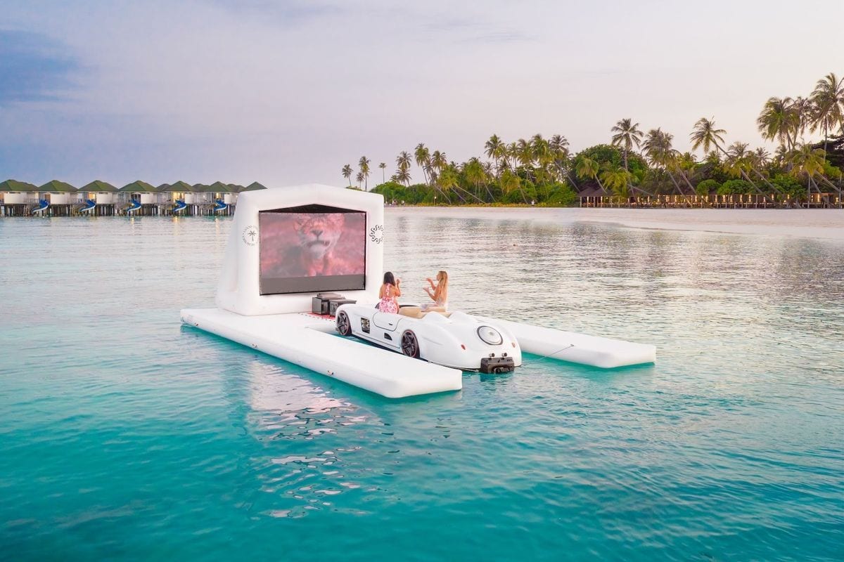 Zwei Personen sitzen in einem weißen, sportwagenähnlichen Boot vor einer aufblasbaren Kinoleinwand, die auf einer schwimmenden Plattform im Meer steht.