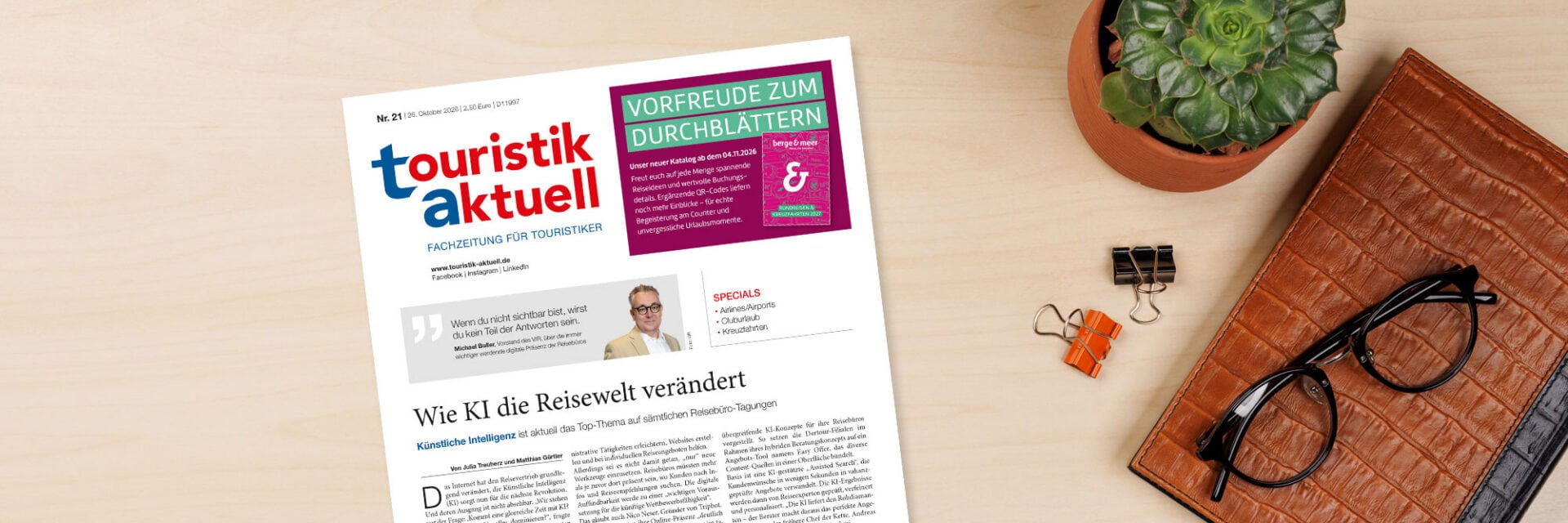 ta-header-21-26_Roman-Valiev_2000px Die Titelseite der Fachzeitung touristik aktuell liegt auf einem Holztisch neben einer Sukkulente, zwei Briefklammern und einer braunen Ledermappe mit Brille.