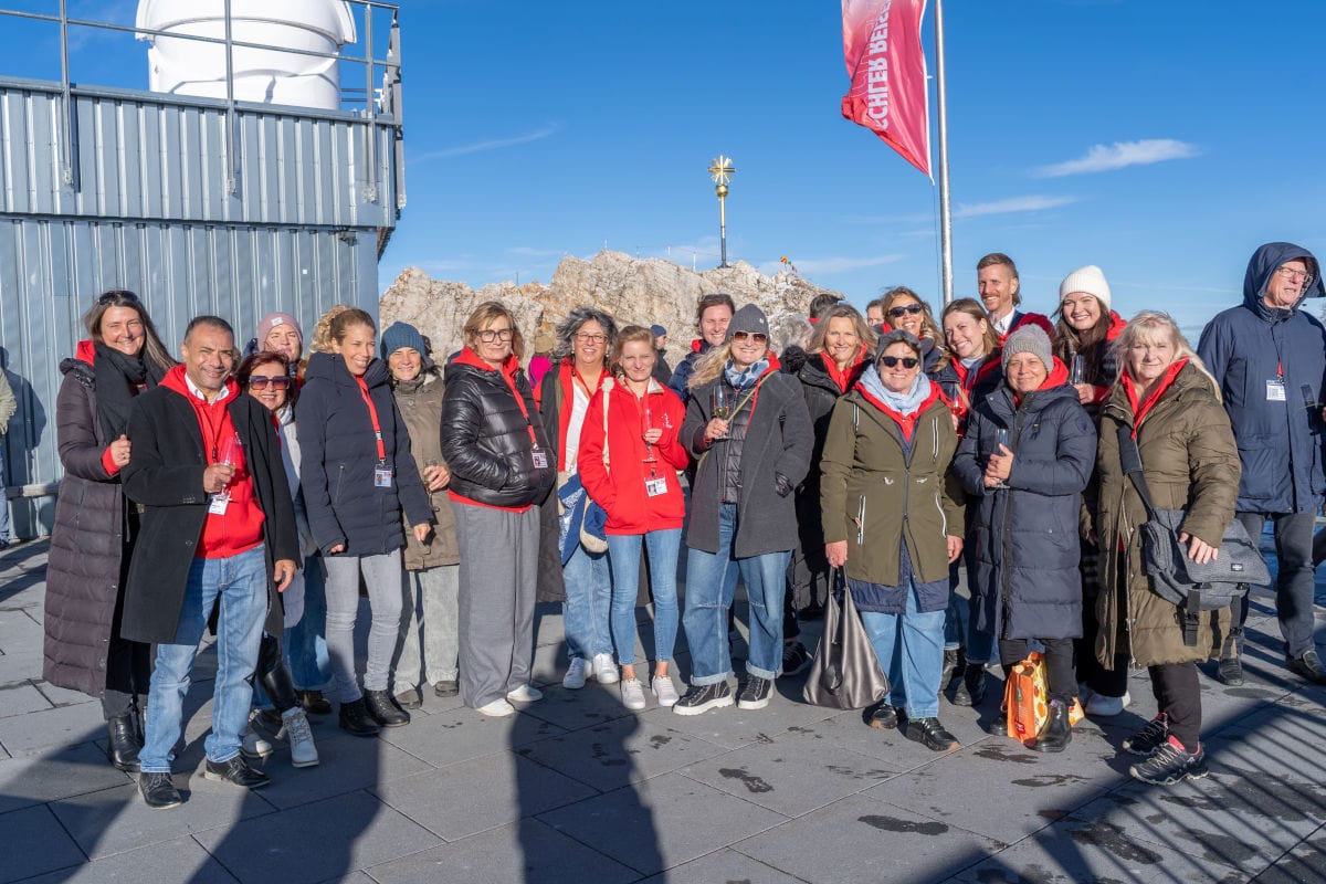 Eine große Gruppe von Menschen steht bei strahlendem Sonnenschein auf einer Aussichtsplattform vor einer Felsformation und einem Gebäude in den Alpen.
