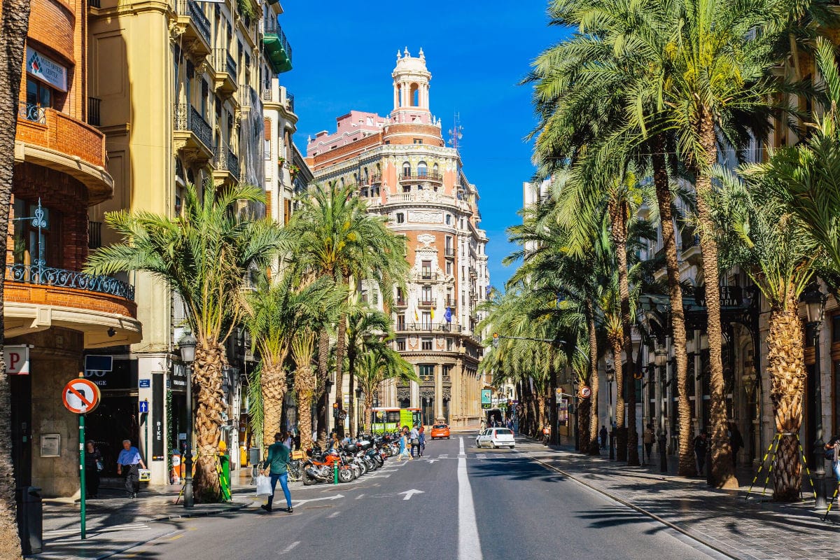 Eine sonnige Straße in Valencia mit Palmenreihen, historischen Gebäuden, parkenden Motorrädern und Passanten unter einem strahlend blauen Himmel.